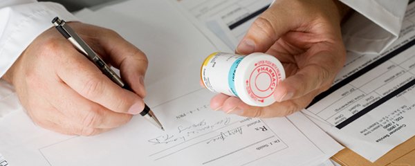 A close-up of a male doctor's hands writing a prescription while inspecting the label of a bottle of medication, illustrating how behavioural science can impact healthcare provider behaviour