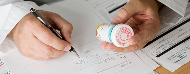 A close-up of a male doctor's hands writing a prescription while inspecting the label of a bottle of medication, illustrating how behavioural science can impact healthcare provider behaviour
